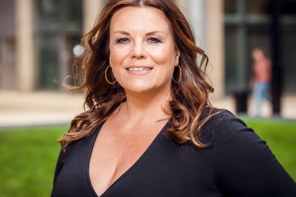 A portrait of Natalie Rogers. Natalie is a white woman with long brown hair wearing a v-necked black top. There is a lawn and buildings in the background